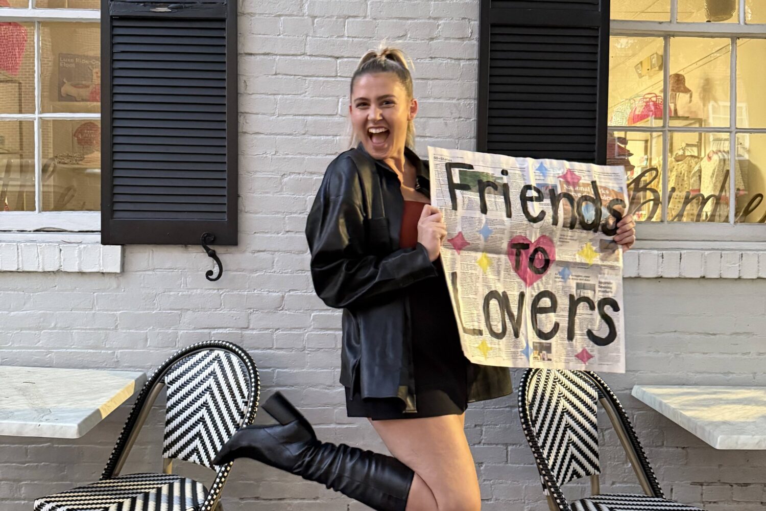 A blonde woman wearing a black coat and boots smiles and holds up a sign that says "Friends to Lovers."